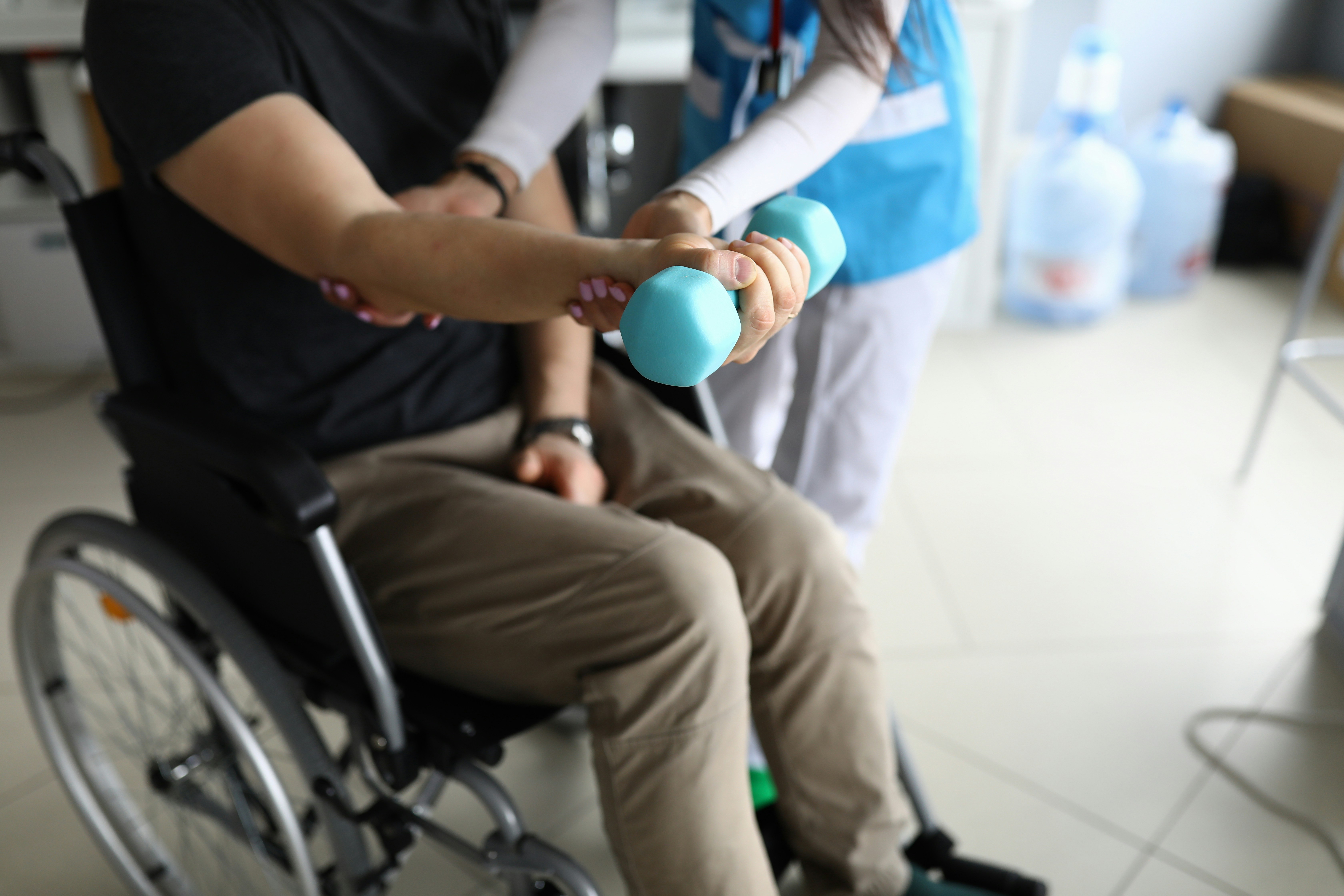 Person using a dumbbell during a rehabilitation session while seated in a wheelchair with support from a healthcare professional.