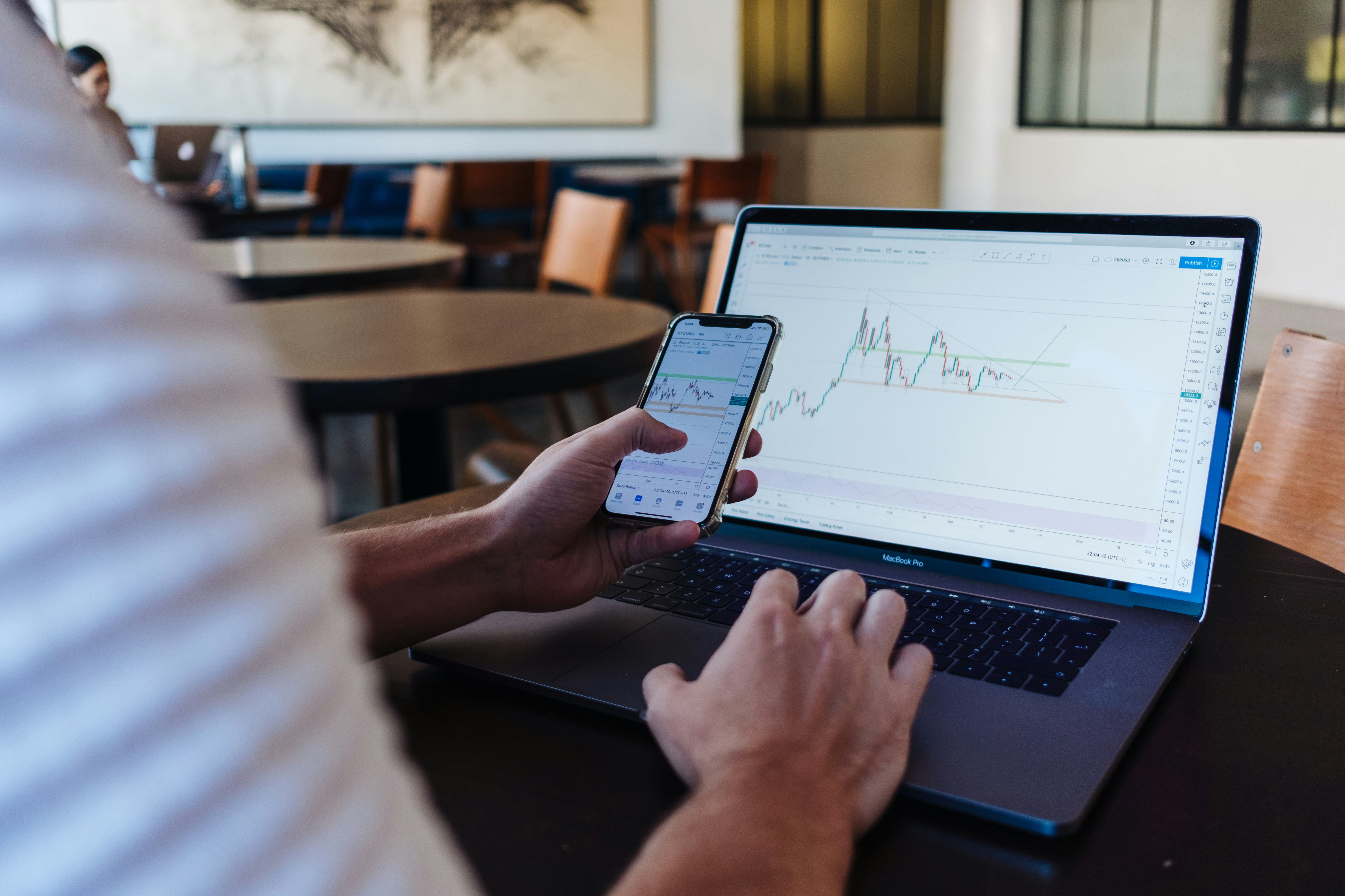 Person using a smartphone and laptop to view cryptocurrency trading charts and digital asset market data in a modern office setting.