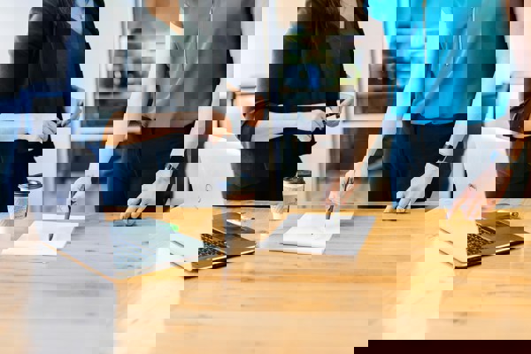 Two people standing at a wooden table in an office setting, with a laptop, a takeaway coffee cup, and a notepad on the table. One person is holding a pen over the notepad, suggesting a discussion or planning session.