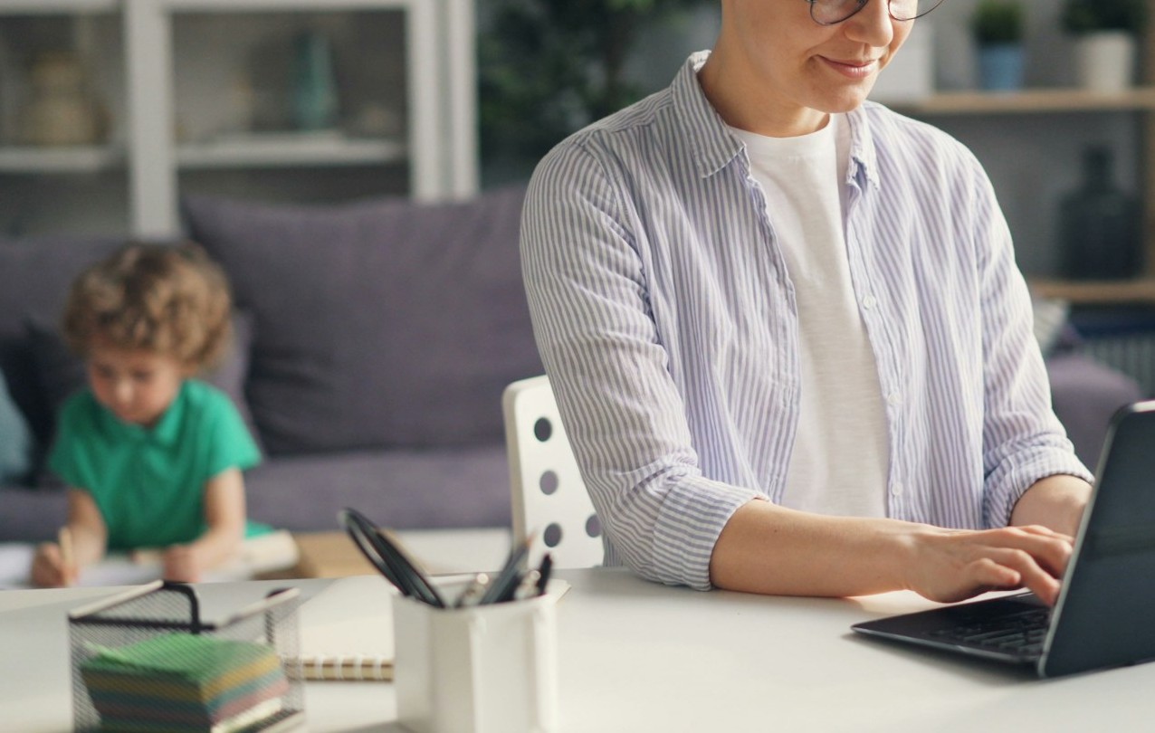 Person working from home on a laptop while caring for a child, representing the impact of caregiving responsibilities on employment and earning capacity.